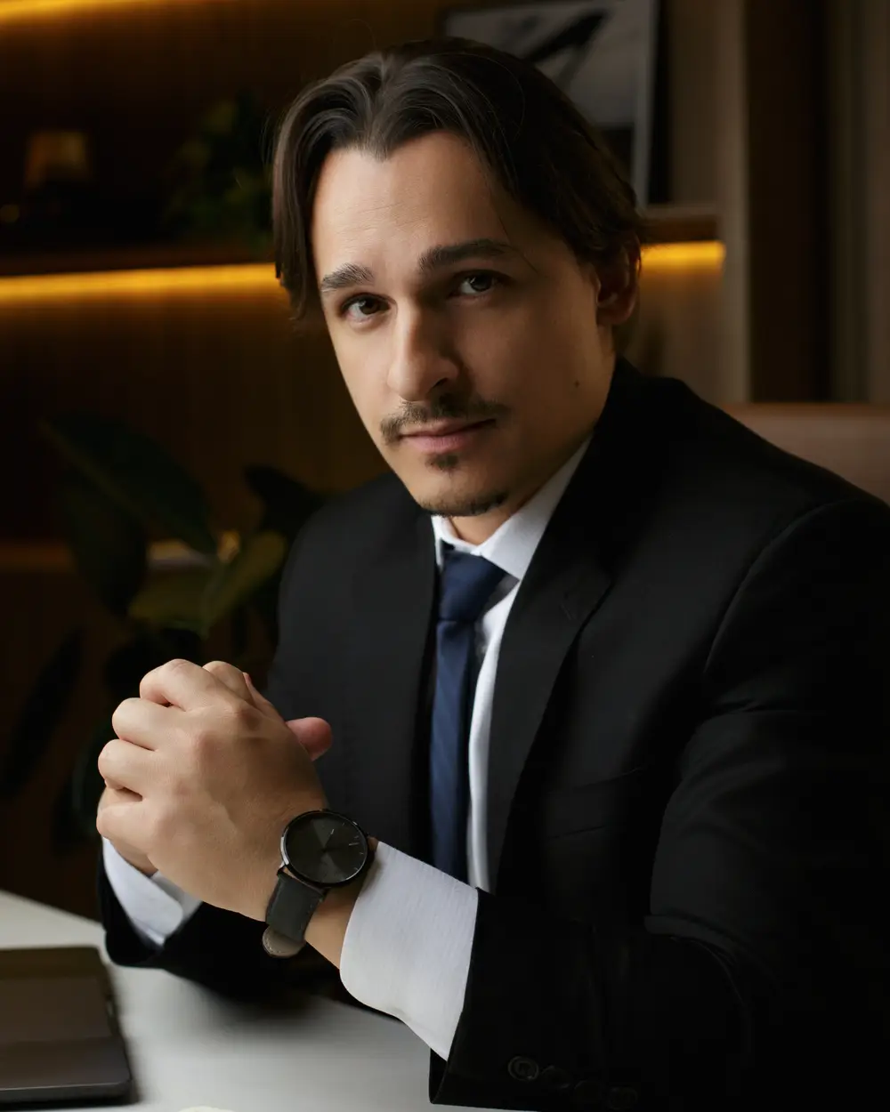 Rafael Dantas sentado à mesa do escritório, de terno, olhando para a câmera